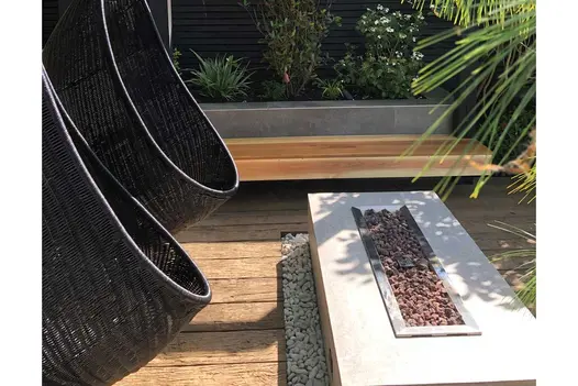 Two black egg chairs hang over vintage millboard decking next to floating bench and firepit, surrounding tropical plants.