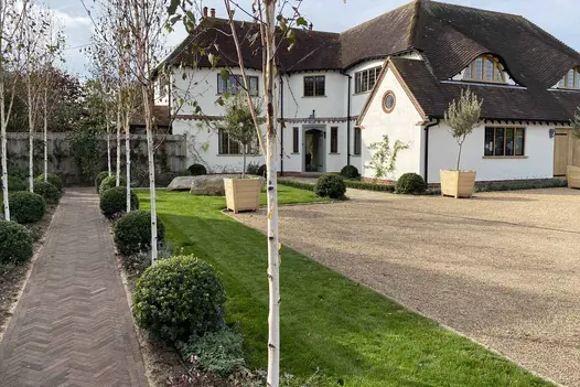 White L-shaped house with 2 eyebrow dormer windows and large gravel drive. Straight Derby brick paving path by long lawn to left.