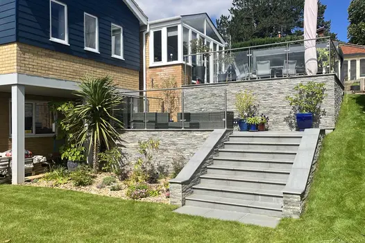 Glass railings edge 2 terraces descending past back of house on steep slope. Kandla Grey sandstone cladding on retaining walls.