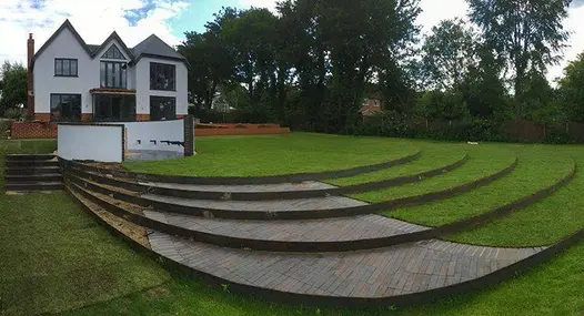 Back lawn descends in 4 wide curved steps, part grassed, part laid with Chelsea paving bricks. White house in background. 