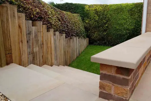 Steps descend between fence of upright wooden sleepers and low parapet topped with Buff swimming pool coping stones.