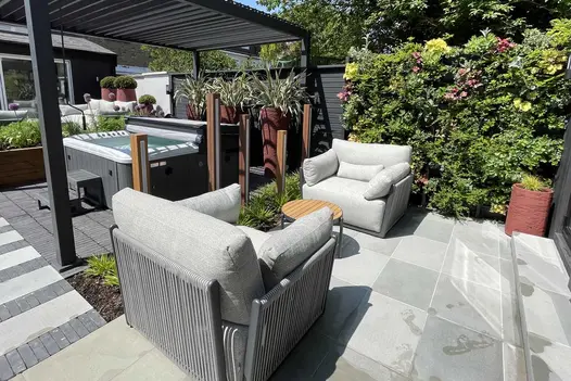 Living wall on fence, behind 2 squashy armchairs on Dove Grey sawn sandstone paving next to pergola over half-open hot tub.