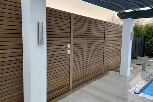 Tall closed-slatted fence of Warm Teak composite battens edges garden with porcelain paving and white posts holding pergola roof.