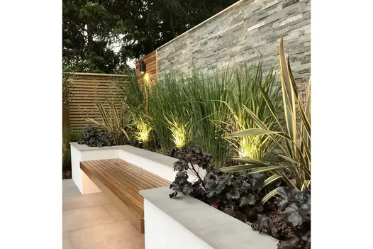 Wooden bench set into white raised bed with spotlights highlighting tall grasses in front of wall of Graphite sandstone cladding.