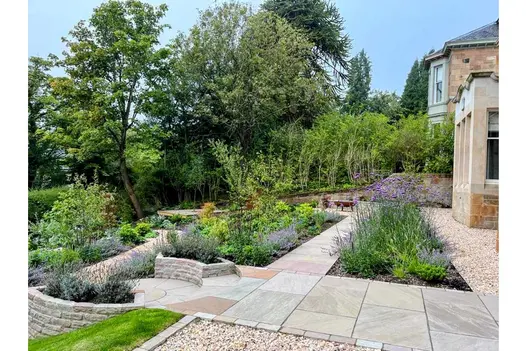 2 curved raised beds of Raj Green sandstone walling embrace paving circle on terrace descending from back of stone-built house. 