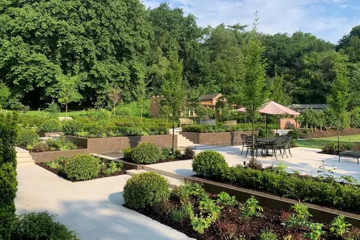 Large patio area with multiple levels, trees border the background. Patio split by large cladd planters filled with greenery.
