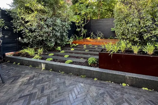 Moderna brick paving in front of step up to staggered path of plank paving leading to wooden bench with black fence behind.