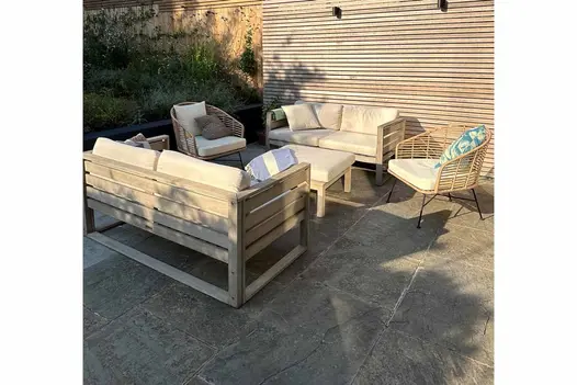 Wooden, cushioned outdoor sofa set in sunny spot on Kandla Grey Indian sandstone paving next to panel of battens attached to fence.