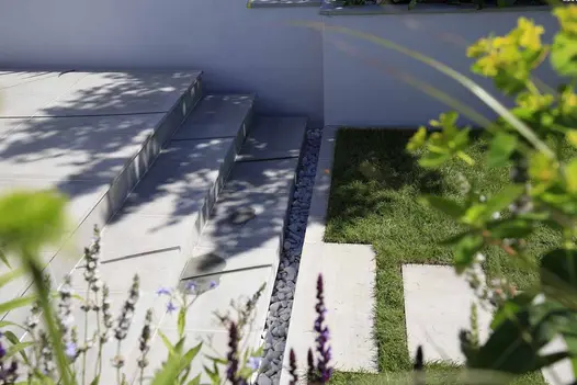 3 steps of Dove Grey sawn sandstone, with 2 widely spaced curved grooves cut into them rise from lawn to paving past white wall.