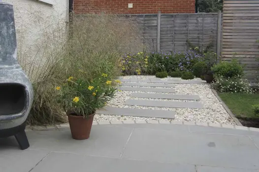 Curve-edged Flamed Grey Smooth Sandstone patio, with plant pot and chiminea, next to gravel path with matching plank format paving.
