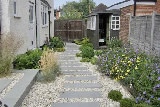Sinuous path of Flamed Grey Smooth Sandstone paving slabs set into gravel runs between house and flower beds to tall wooden gates.
