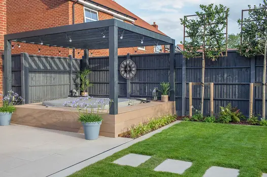 Florence Beige porcelain paving slabs, laid in lawn, lead to matching patio with metal slot drain and raised hot tub under pergola.