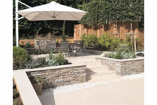A patio elevated with Florence Beige porcelain slabs, adorned with a metal dining set and pergola, encircled by raised planter beds.