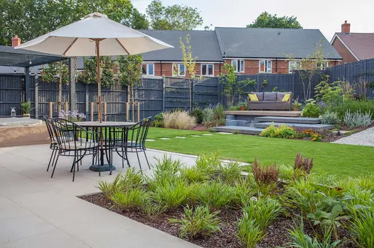 Deep plant border set into Florence Beige porcelain patio, edging lawn where matching paving slabs lead to raised seating area.