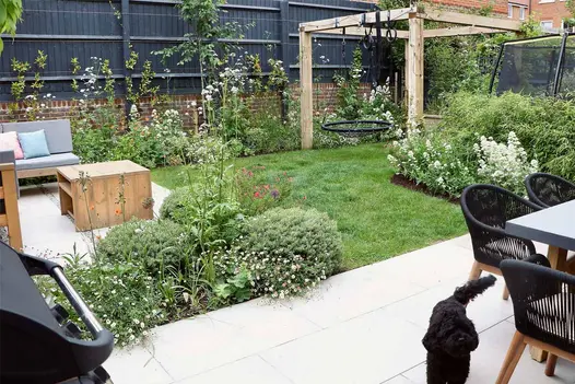 Black dog on florence beige porcelain paving with dining table to the right and lounge set to the left. Lawn in between with pergola and swing.