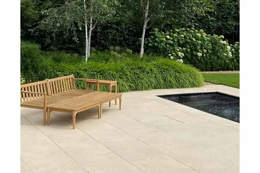 Koi pond in the background, with wooden lounge chairs opposite sat on Florence Beige Porcelain Paving. Lots of greenery surrounding.