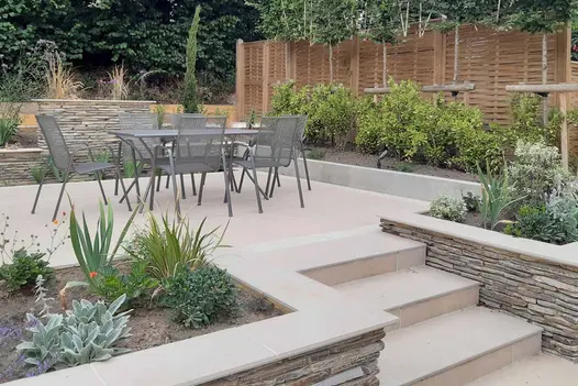 Steps with flowerbeds either side lead up to patio are with dining table using florence beige porcelain paving.