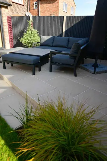 Dark outdoor furniture sits on light florence beige porcelain paving in the shade, sunlight pours over black painted fence.