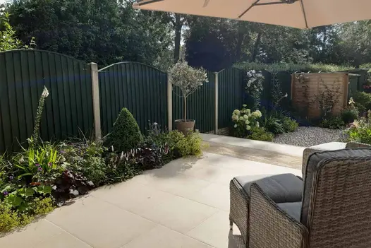 Rattan armchair faces florence beige porcelain patio with flowerbeds against green fencing, steps lead down to pebbled area.