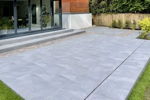 Rectangular patio of cloudy grey Florence Storm outdoor tiles separated from steps to bifold patio doors by clay paver strip.