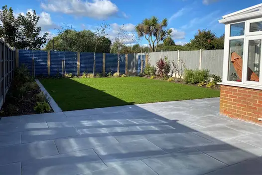 Patio in Florence Storm porcelain paving wrapped around room at rear of house. Matching edging separates wide flower bed from lawn.