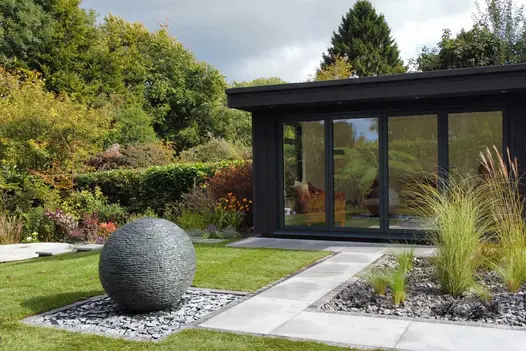 Mature rear garden featuring a modern glass fronted home office, grey porcelain paving paths and a large floating ball water feature.