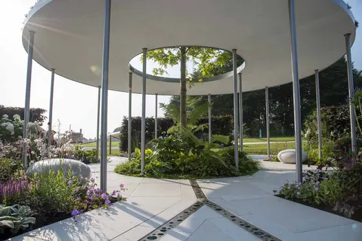 Tree pokes through hole in centre of roof of round pergola above circular flowerbed set in Florence White Porcelain patio tiles.