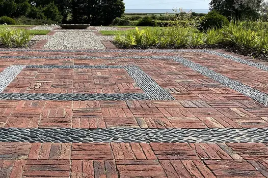Large paved area of Old English Belgian bricks laid basketweave with pebble detailing leads to gravel path to water bowl.
