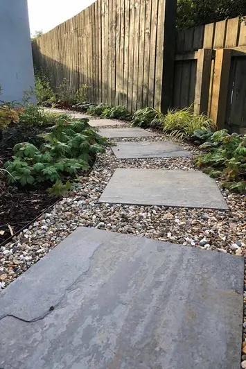 Rustic Slate Paving slabs used as stepping stones curving around path of pebbles. Flowerbeds either side.