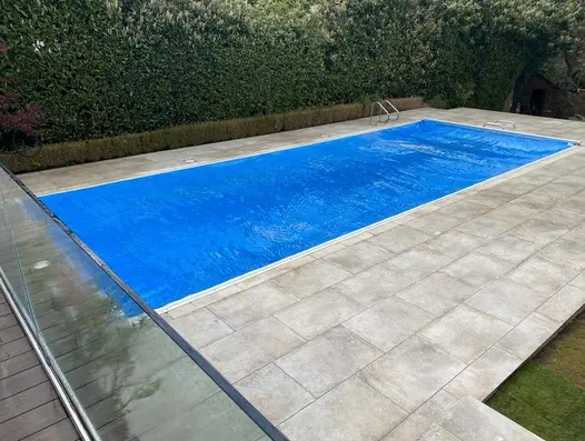 Frosty Grey 16mm Porcelain Paving around outdoor swimming pool, framed with hedging and modern glass balustrade.