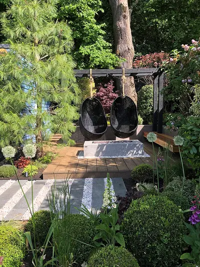 In woodland garden, 2 egg chairs hang over raised deck next to wide striped path of Black Granite plank paving and white pebbles.