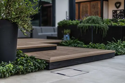 Porcelain paving leads up to steps clad in millboard decking, using burnt cedar fascias, with hedges spilling out from ground and planters.