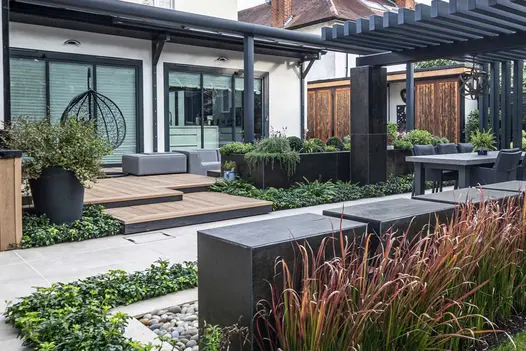 Swinging chair in front of french doors has decking area in golden oak millboard leading down as steps to patio, pergola and dining area.