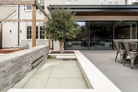 Wide spout in low grey clad wall pours water into rectangular pond with Comblanchien porcelain tiles. Design by Gadsden Gardens.