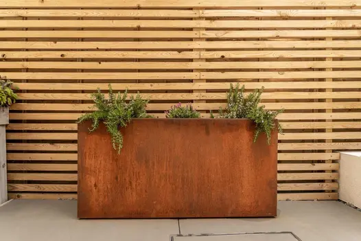 Corten steel tall trough planter stands against high slatted fence, near raised beds with white walls. Dining set in background.