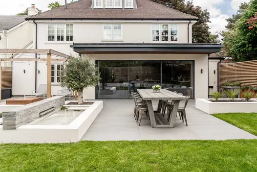 Patio with long table next to raised rectangular pond backed by low wall of Kandla Grey stone cladding. Built by Gadsden Gardens.