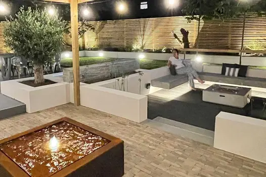 Low wall faced with Kandla Grey Sandstone External Cladding creates feature next to olive tree, sunken seating area, and small lawn.