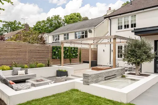Low wall of Kandla Grey sandstone external cladding backs raised pond next to olive tree, adjacent to sunken seating area.