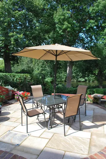 Metal dining set and pergola on round Ming Indian sandstone patio edging with soldier course of brick pavers. Tall trees behind.