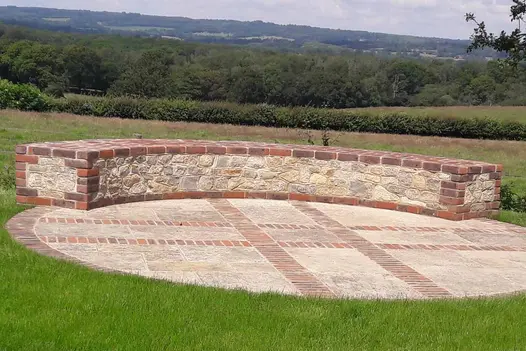 Curved block borders a paved circle in grass on edge of field. Edges and corners are outlined with Old English garden bricks.