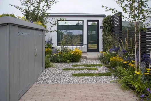 Small, narrow show garden, paved with bricks and gravel, has tall fence of Dark Ash composite battens down one side. Shed on other.