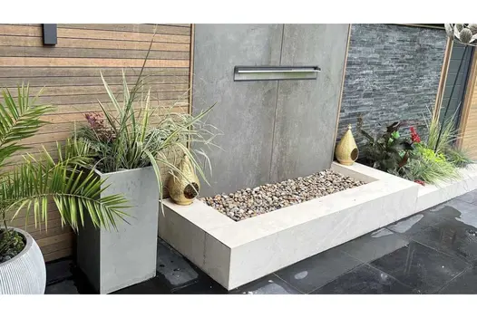 Grey-clad wall with water spout separates stone cladding from Warm Teak composite battens on boundary of paved area with pots.