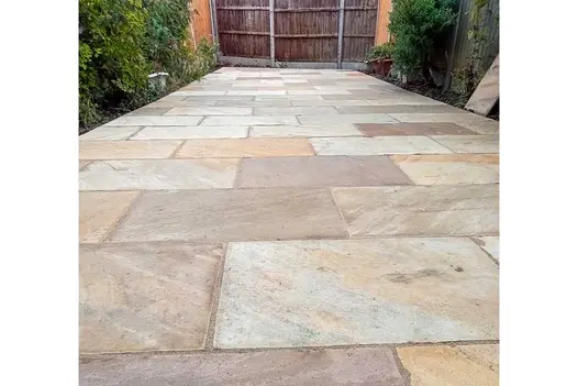 Long rectangular area paved with mint Indian sandstone with tall wooden fence at end. Planted borders on either side.