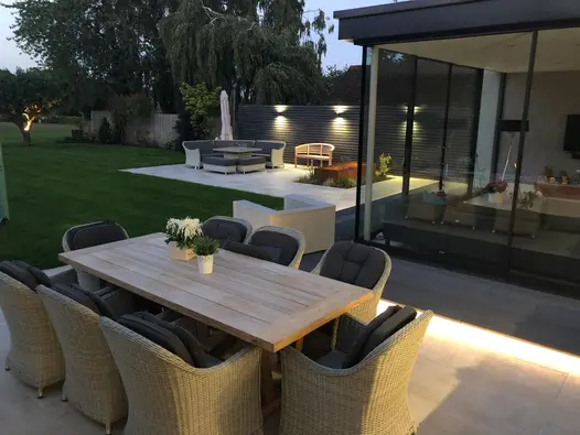 8 seater outdoor dining table adjacent to large glass extension looking down towards another paved seating area and large rural garden.