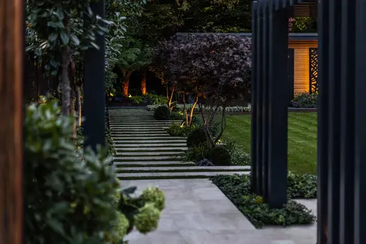 Patio of Cream porcelain paving slabs adjoins stripy path of matching paving and grass next to multi-stem trees and lawn.