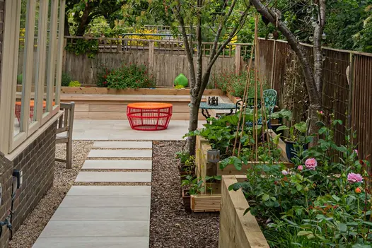 Cream porcelain paved path set in gravel leading towards a patio with built in seating and raised new sleeper flower beds.