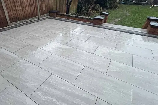Large square raised patio of wet Kandla Grey porcelain paving and low brick edge sits above grassed garden with mature shrubs.