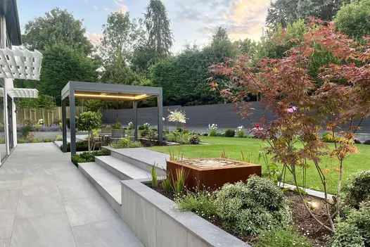 multi level patio featuring proteus pergola uses platinum grey porcelain paving for garden tiles and steps leading up to lawn.