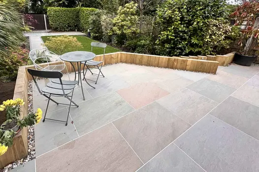 Variation of colour shown in large Raj Green Porcelain Paving patio bordered by wooden planters, hedges, trees and small lawn in the background.