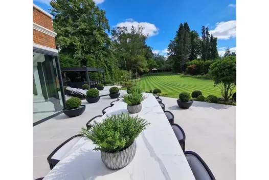 Very long table with chairs on large patio of Urban Grey Porcelain patio tiles. Huge striped lawn and mature trees in background.